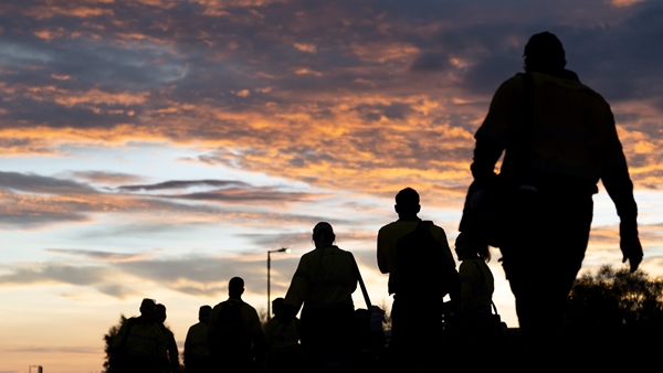 Silhouettes of several people wearing high-visibility work clothing walk together outdoors at dawn or dusk, carrying bags and equipment against a sky filled with orange and purple clouds, with a streetlight visible in the background.